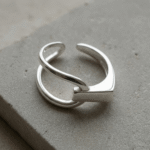 Silver-colored geometric open ring on a white background, showing crossing curved lines, a broad rectangular front, and separated ends.