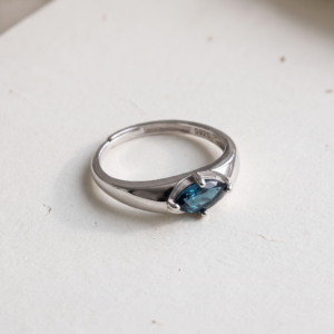 Front view of a marquise london blue topaz ring on a white background, showing the horizontal deep blue-green gemstone, four-prong setting, and overlapping silver-colored band.