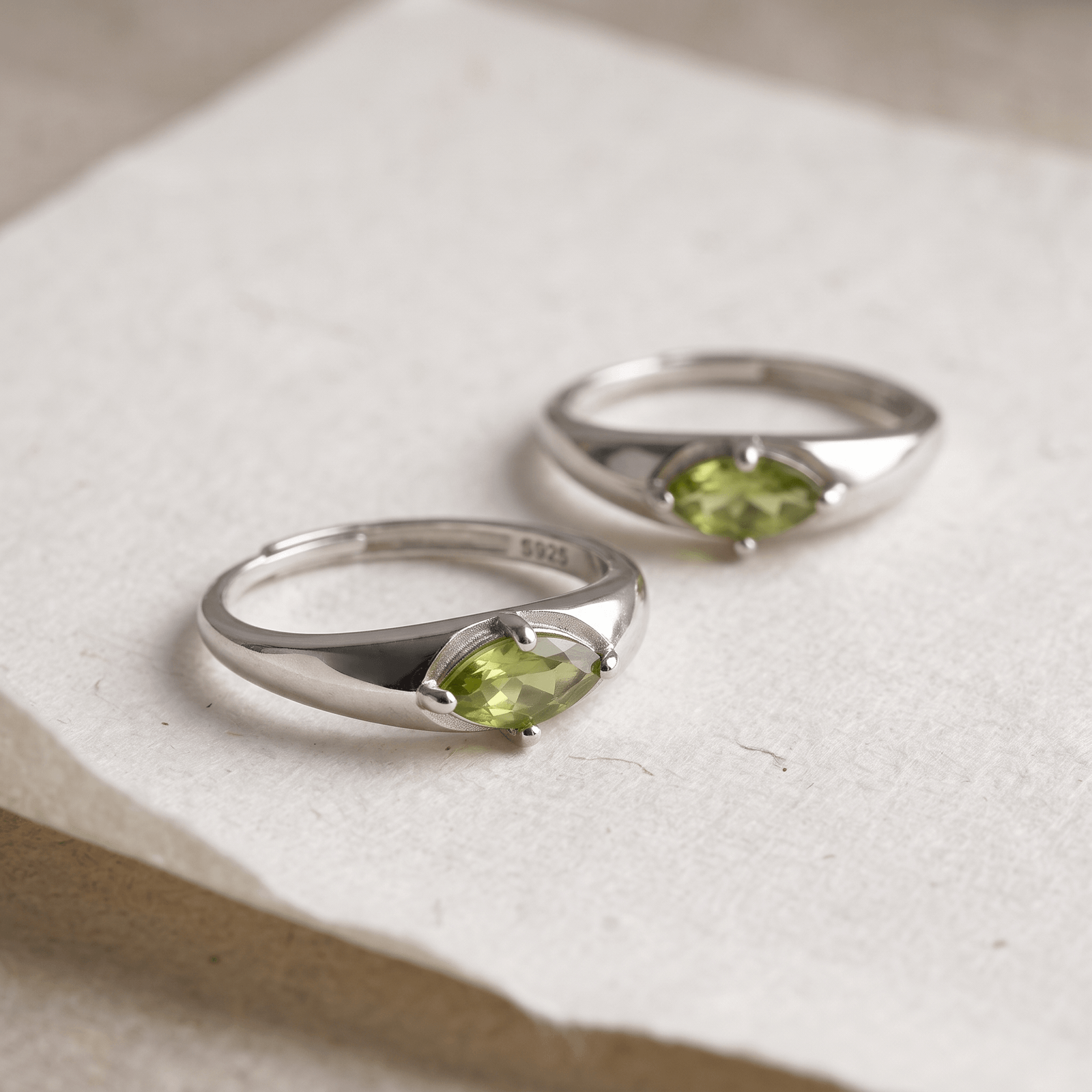 Two silver-colored marquise peridot rings on textured white paper, with the foreground ring in sharp focus.
