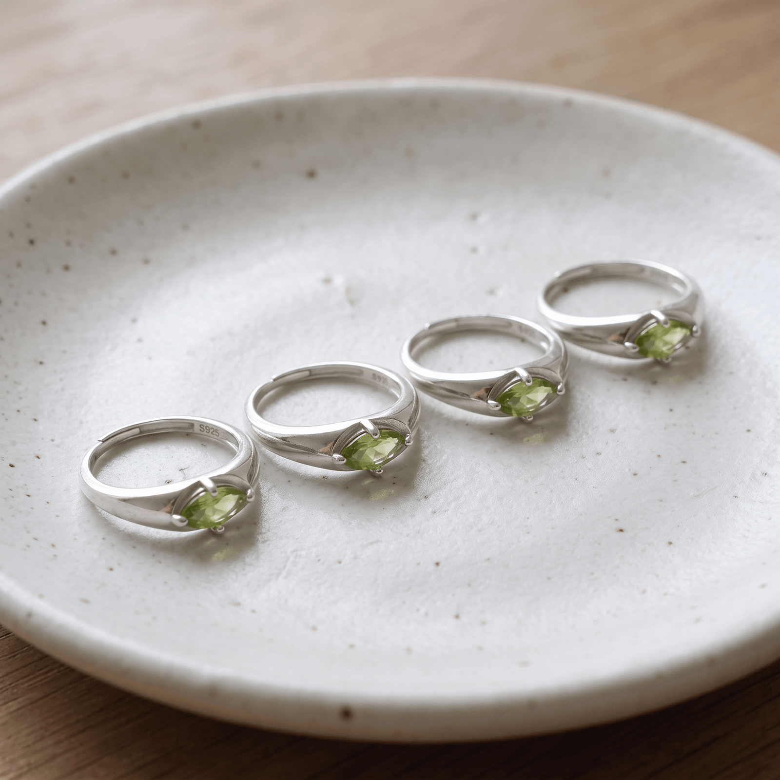 Four silver-colored marquise peridot rings arranged diagonally across a white speckled dish.