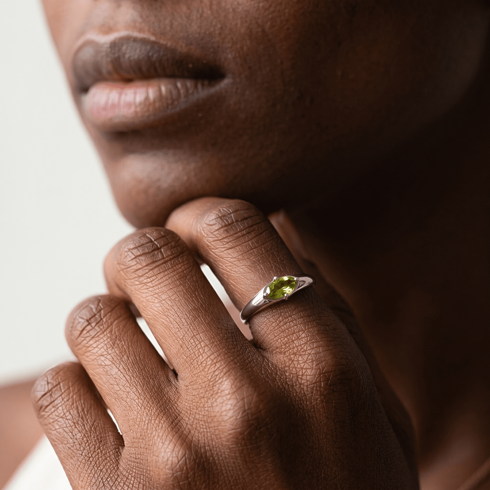 Close-up of a marquise peridot ring on a hand resting beneath the chin, highlighting the green facets and silver-colored band.