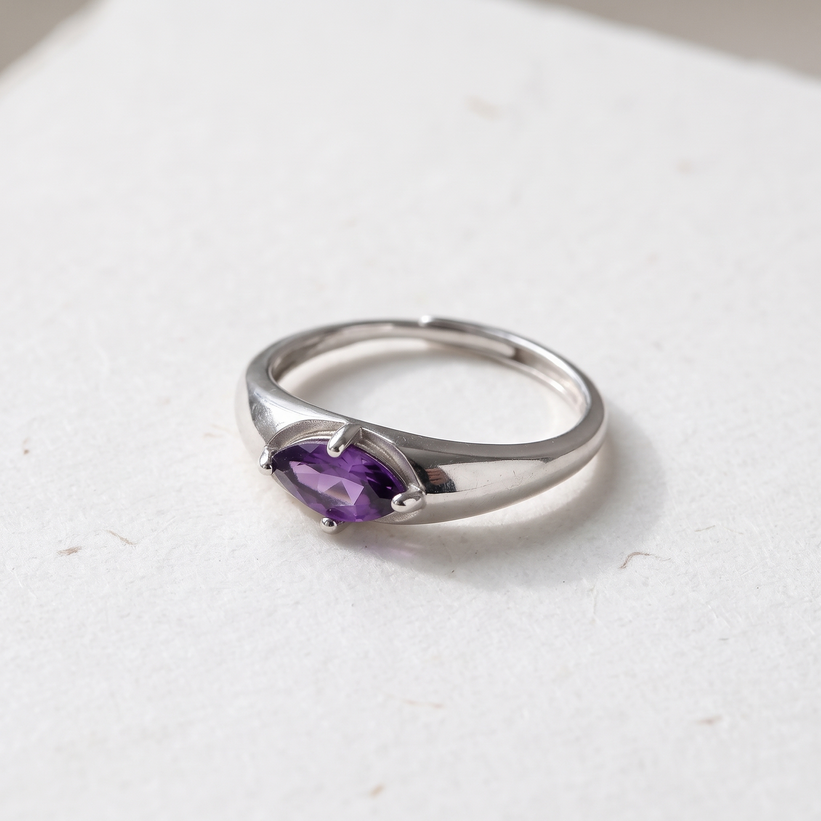 Close-up of an amethyst ring on cream textured paper, highlighting the purple facets, four prongs, and overlapping open band.