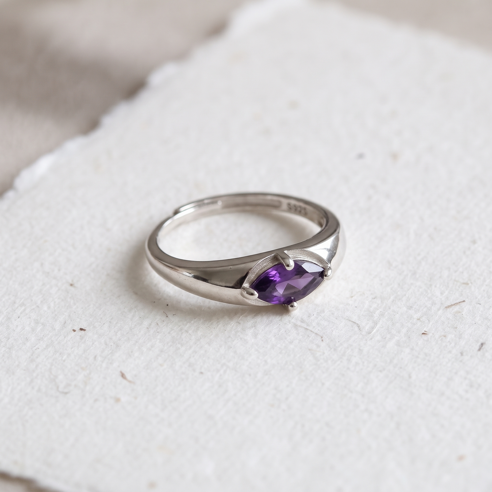 Angled view of an amethyst ring on textured white paper, showing the purple gemstone, four-prong setting, and tapered silver-colored shoulders.