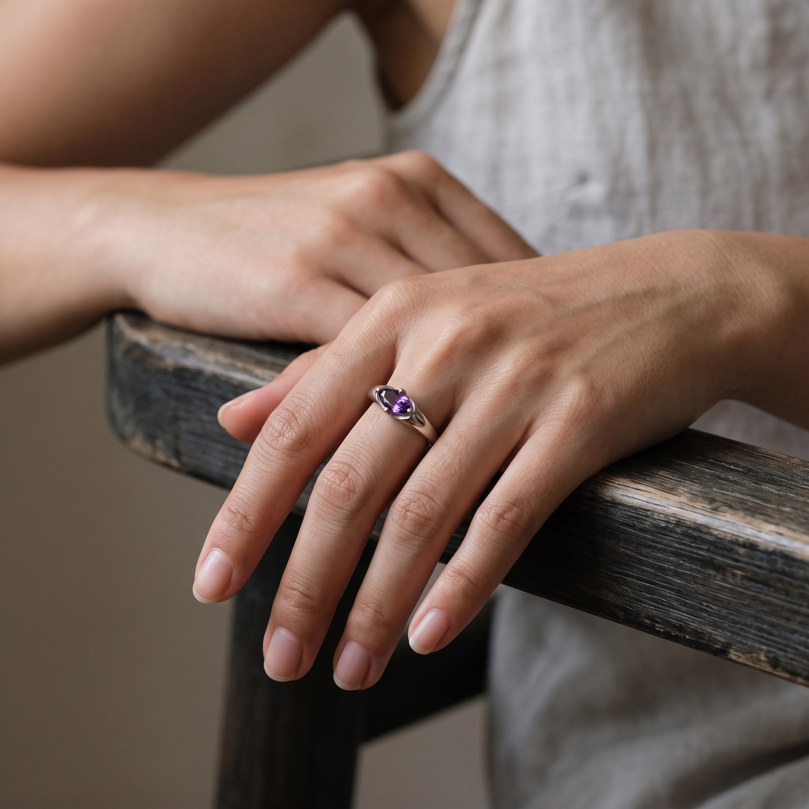 Amethyst ring worn on a relaxed hand resting over a dark wooden chair arm, with a light sleeveless outfit in the background.