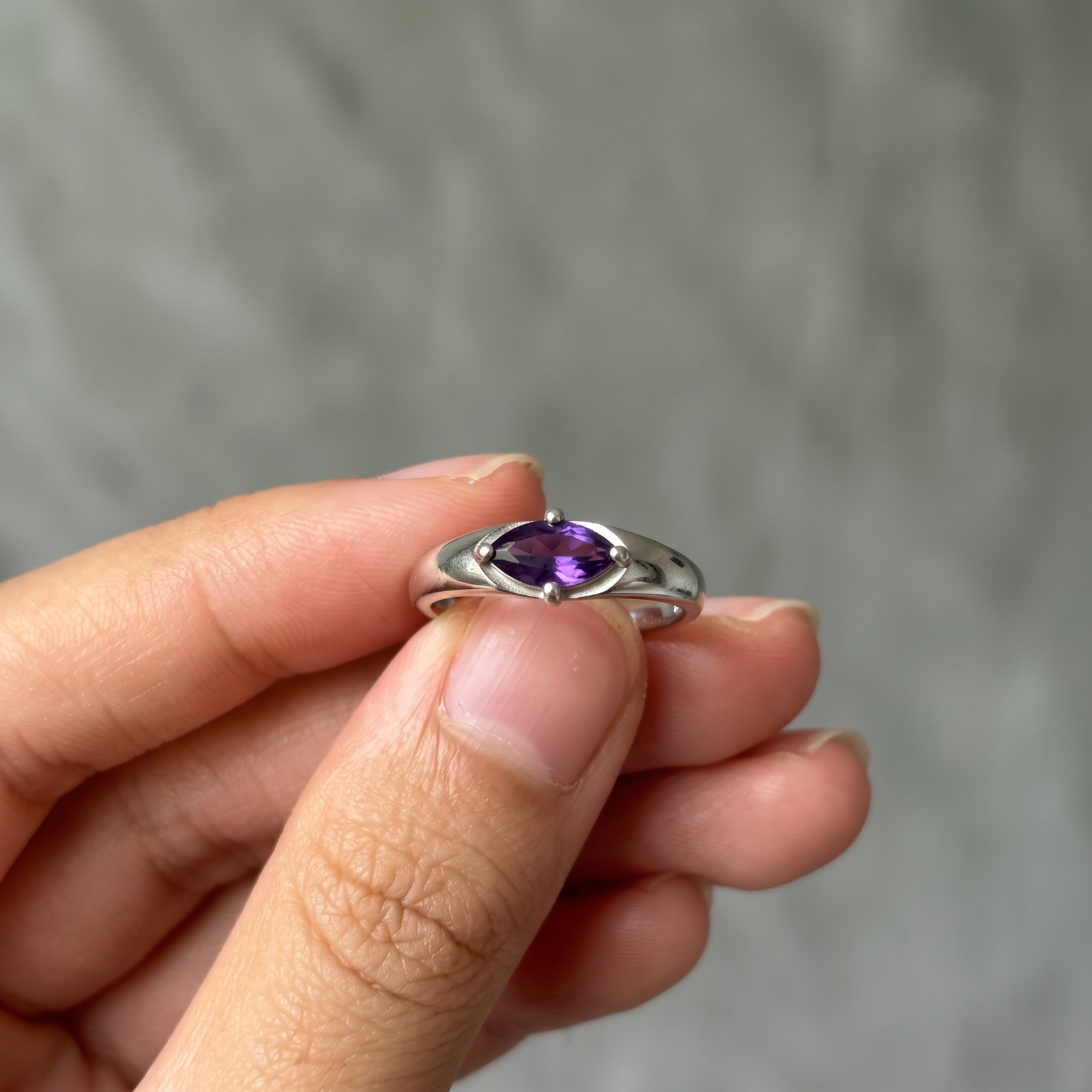 Amethyst ring held between fingertips against a gray background, with the horizontal purple gemstone and four prongs facing forward.