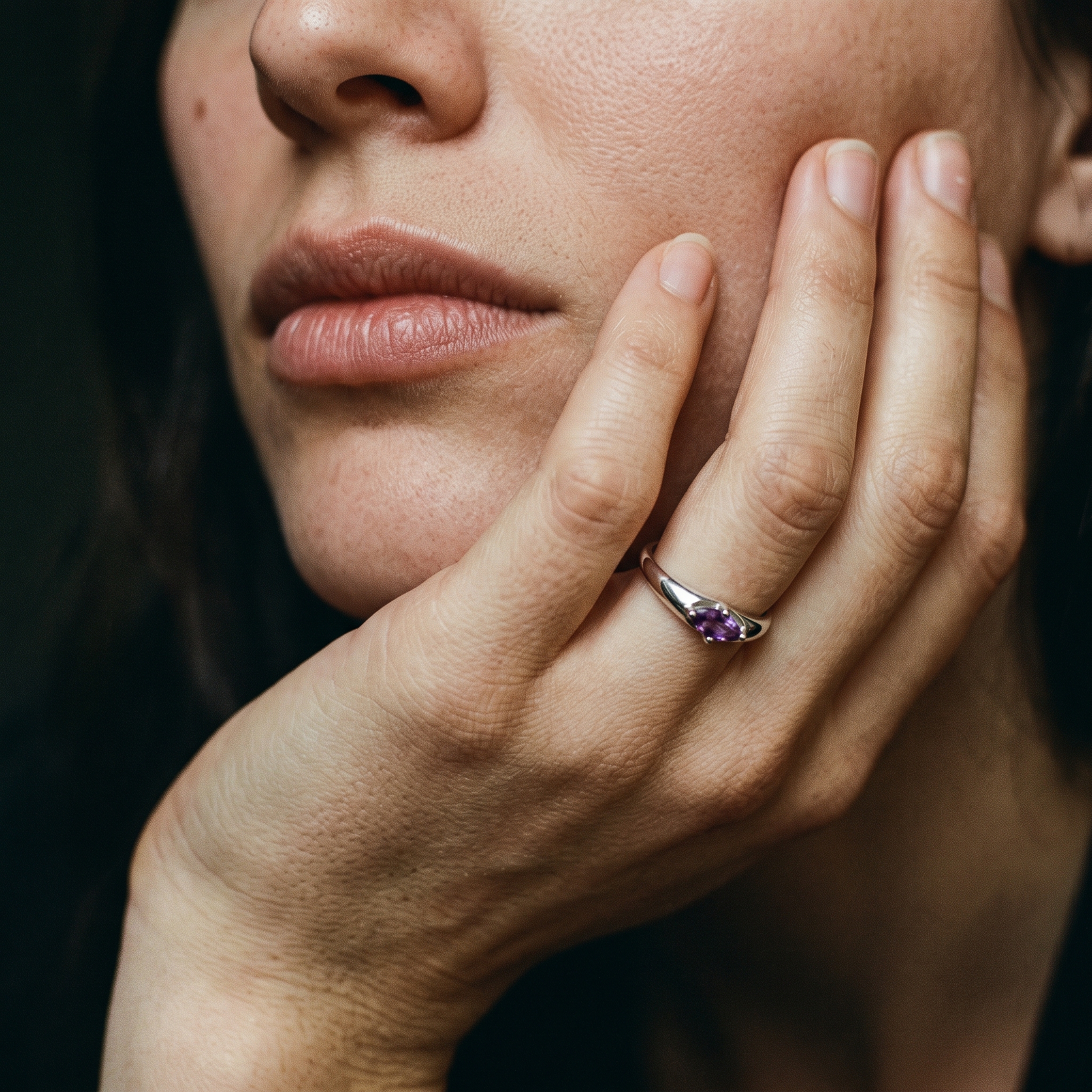 Amethyst ring worn on a hand cupping the cheek in soft, subdued light against a dark background.