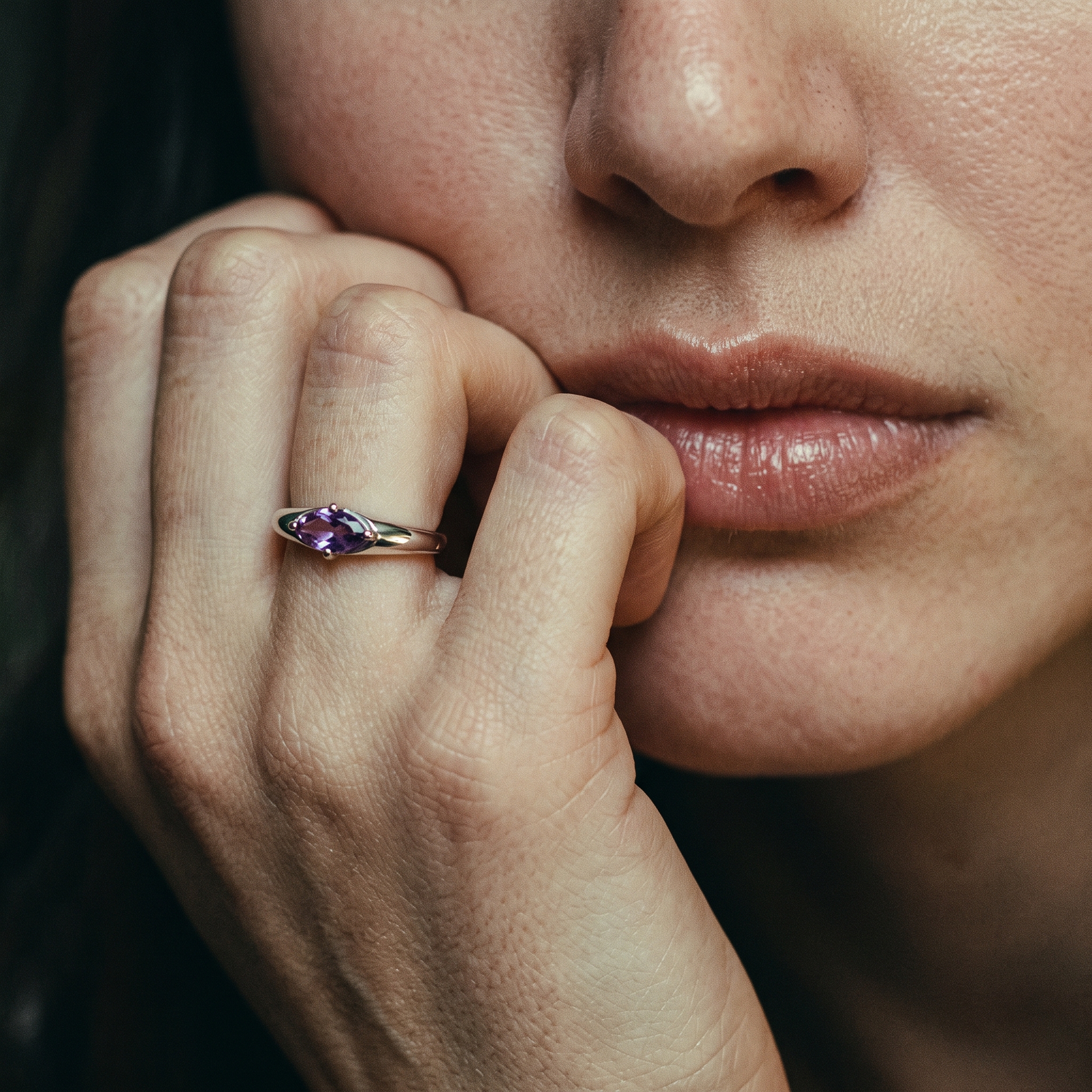 Close-up of an amethyst ring on curled fingers beside the lips, showing the horizontal purple stone and reflective silver-colored band.