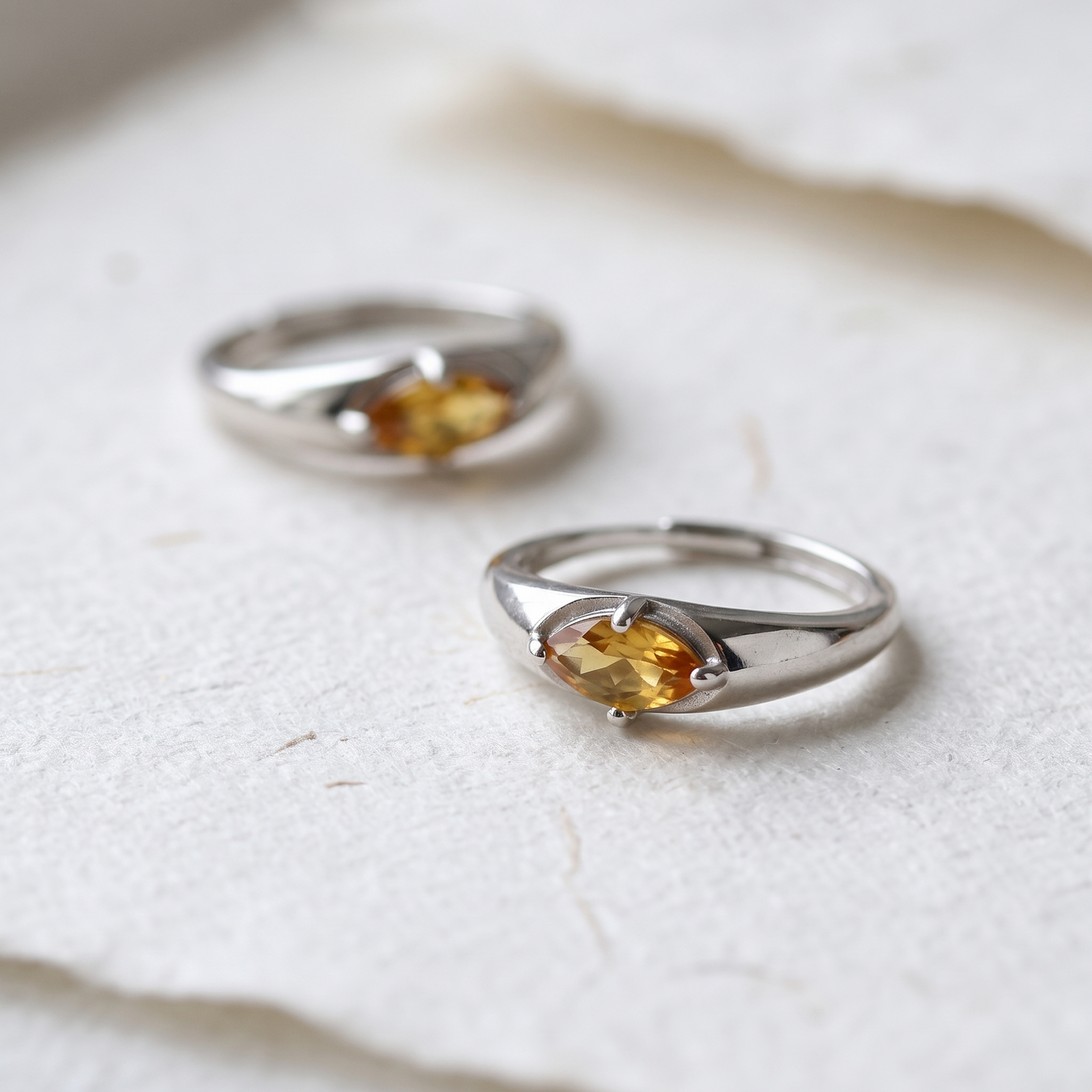 Two silver-colored citrine rings on textured white paper, with the foreground gemstone and setting in focus.