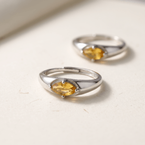 Close-up of a marquise citrine ring on cream textured paper, showing its golden-yellow facets and four-prong setting, with a second ring blurred behind.