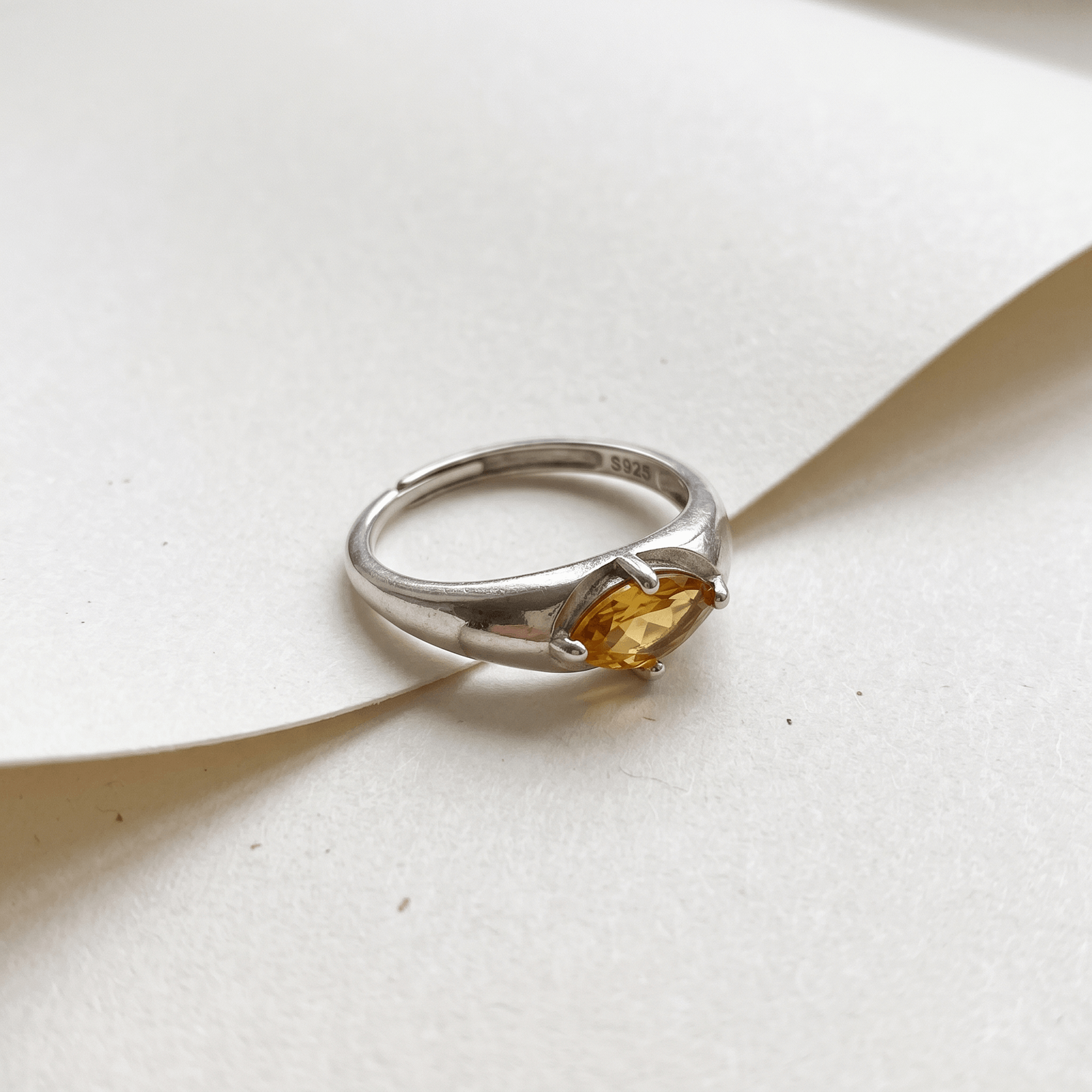 Silver-colored citrine ring resting across overlapping sheets of cream paper, with the faceted golden-yellow stone facing forward.